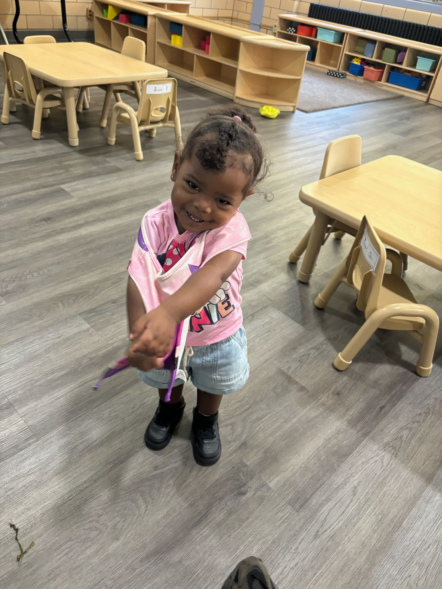 A little girl is standing in a classroom with tables and chairs.