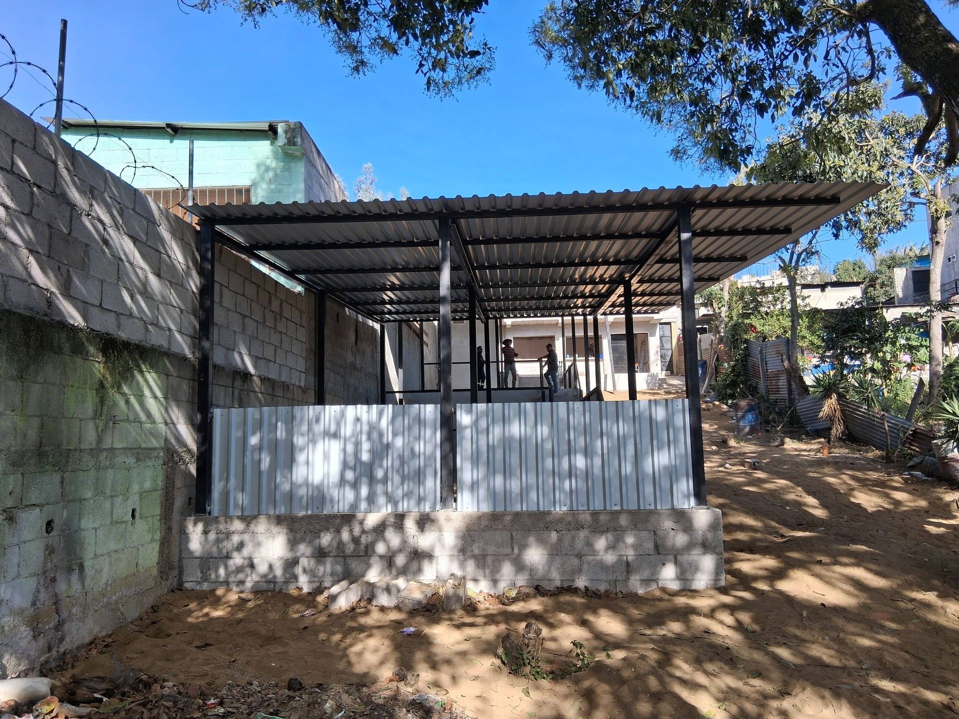 Metal-framed shelter with corrugated roof and walls, constructed near a concrete and brick wall under a blue sky.