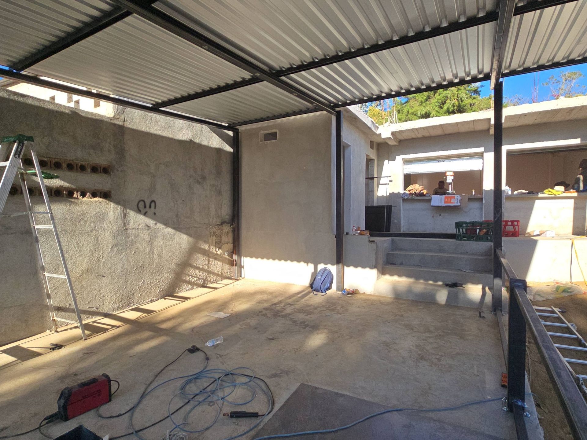 Construction site, covered area. Metal frame supports corrugated roof. Walls are concrete. Tools and ladder visible.