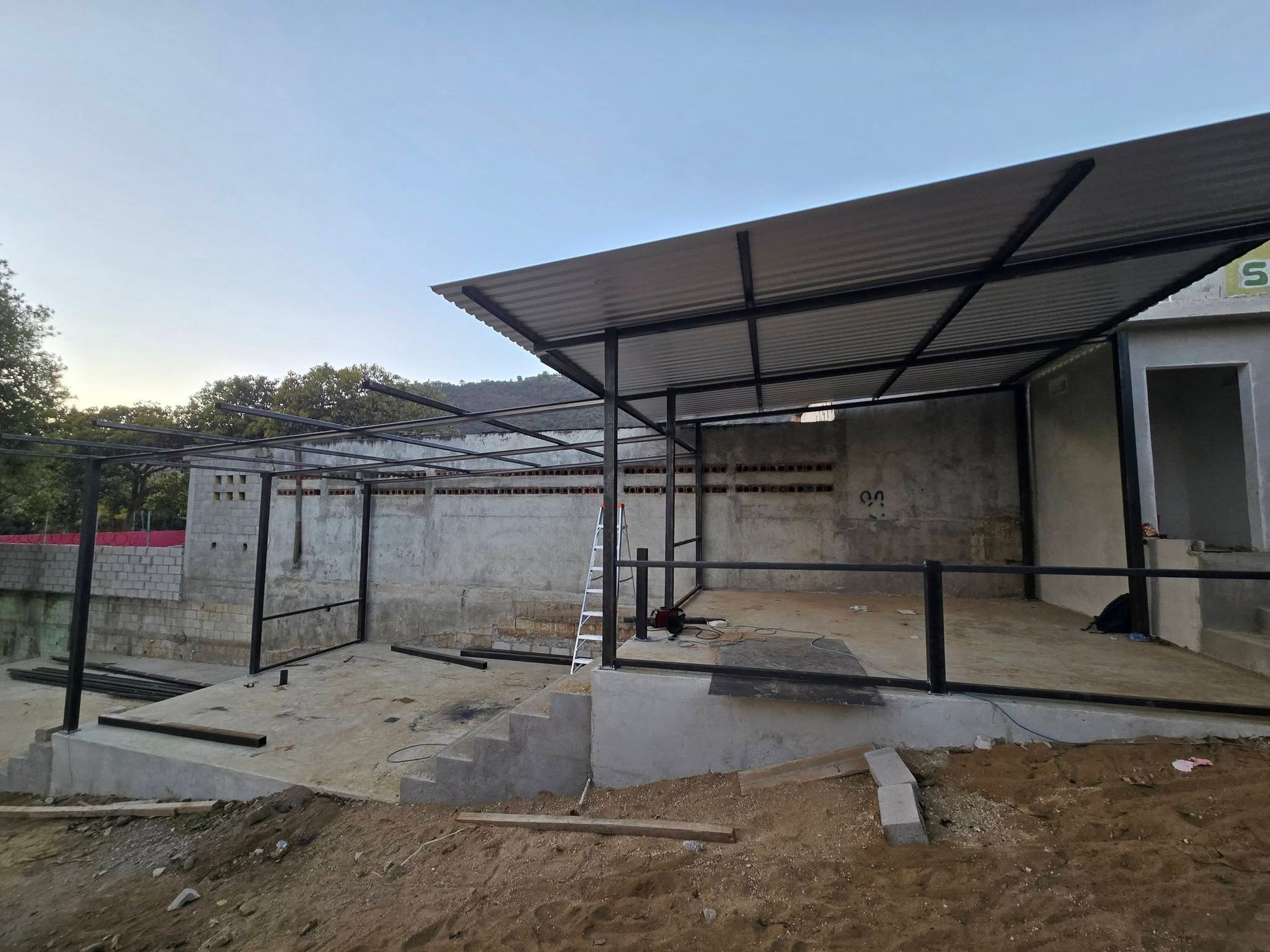 Steel-framed shelter with corrugated metal roof. Concrete walls and ground, construction site.