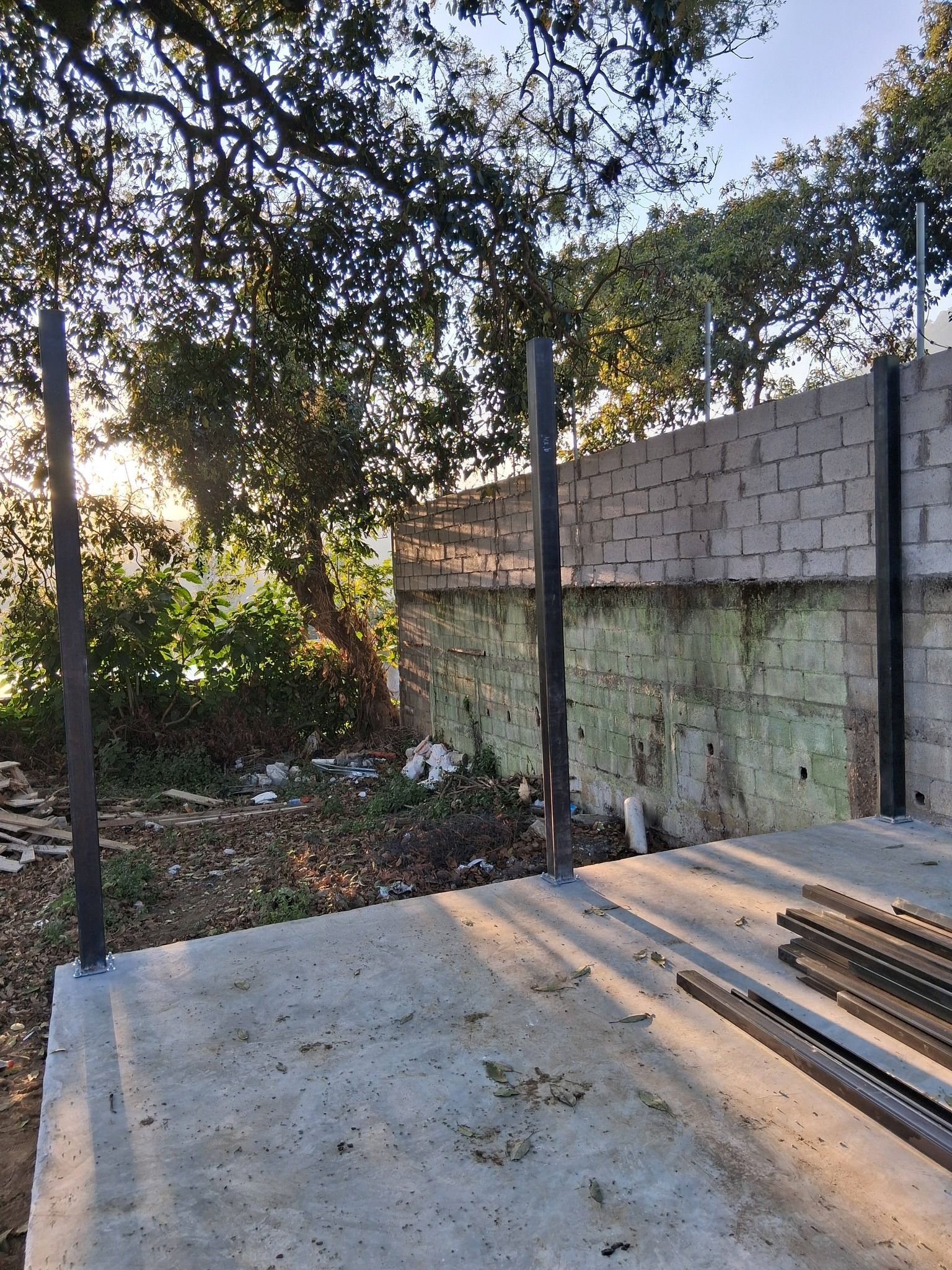 Construction site: metal poles set in concrete slab, brick wall in background.