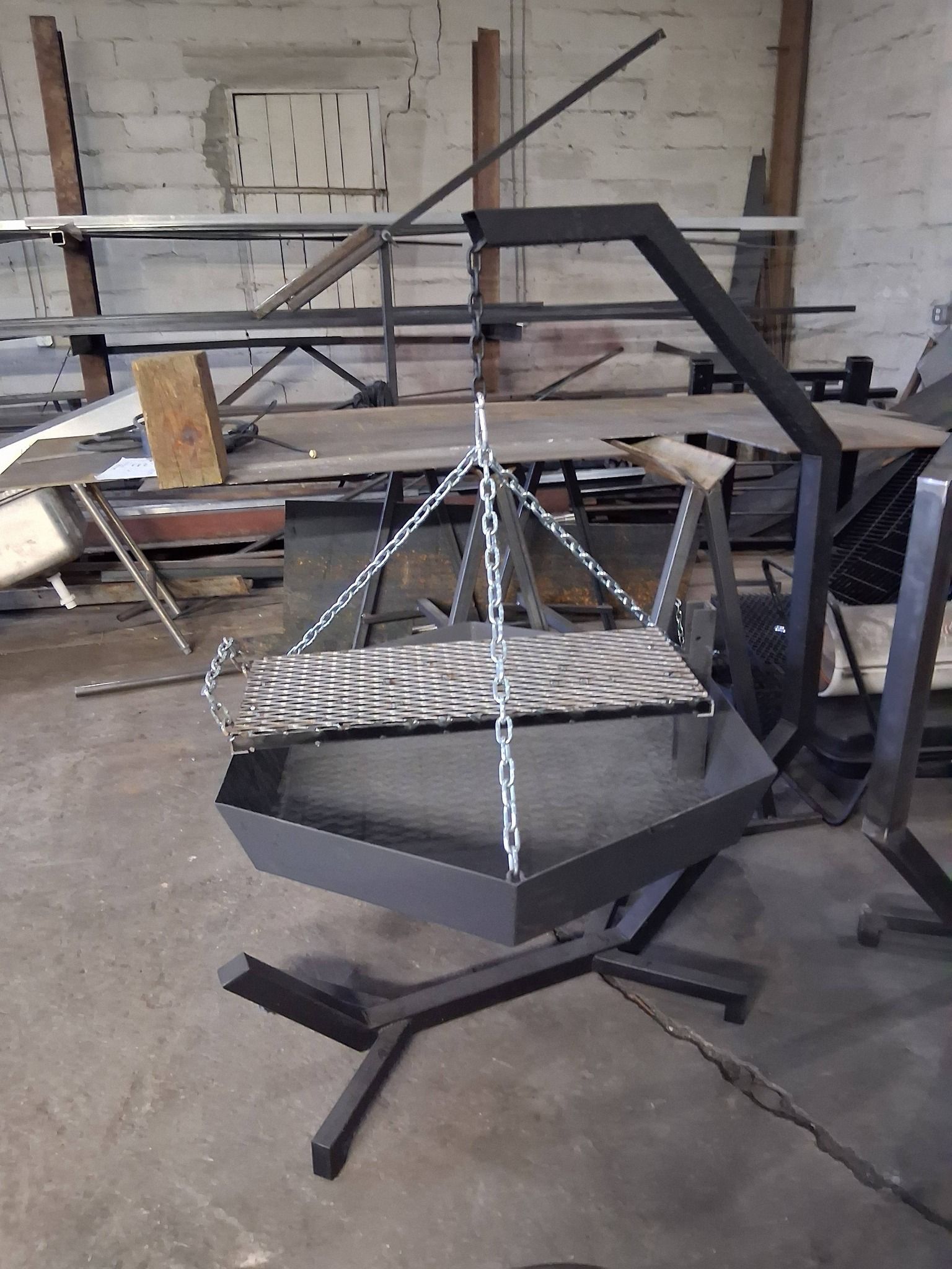 Hexagonal metal fire pit hanging from a black metal frame in a workshop.