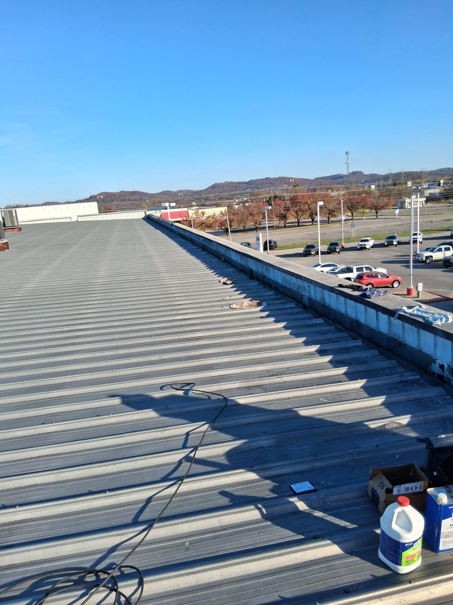 View from a large rooftop overlooking a parking lot, buildings, and hills under a clear blue sky.