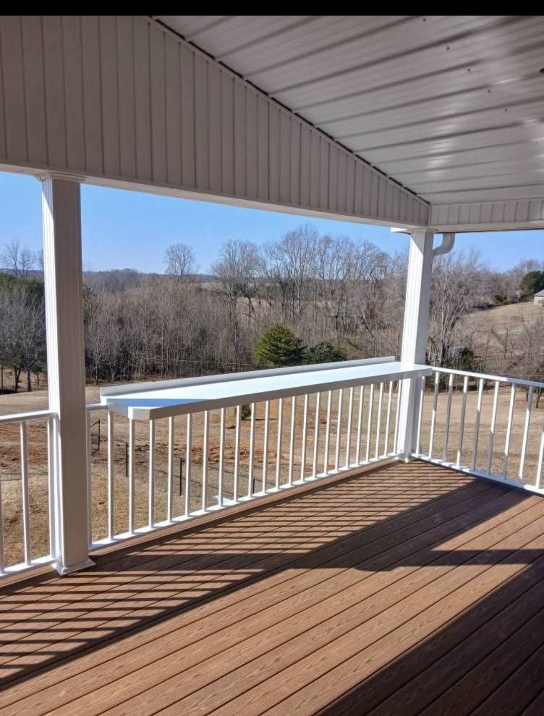 A white porch with brown decking, a railing, and a counter overlooking a field and trees on a sunny day.
