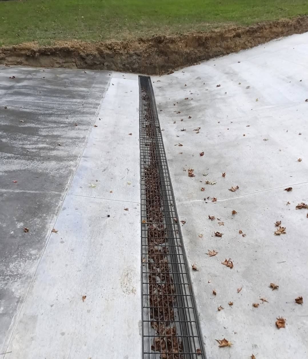 Concrete driveway with a central drainage channel filled with leaves. Green grass borders the driveway.