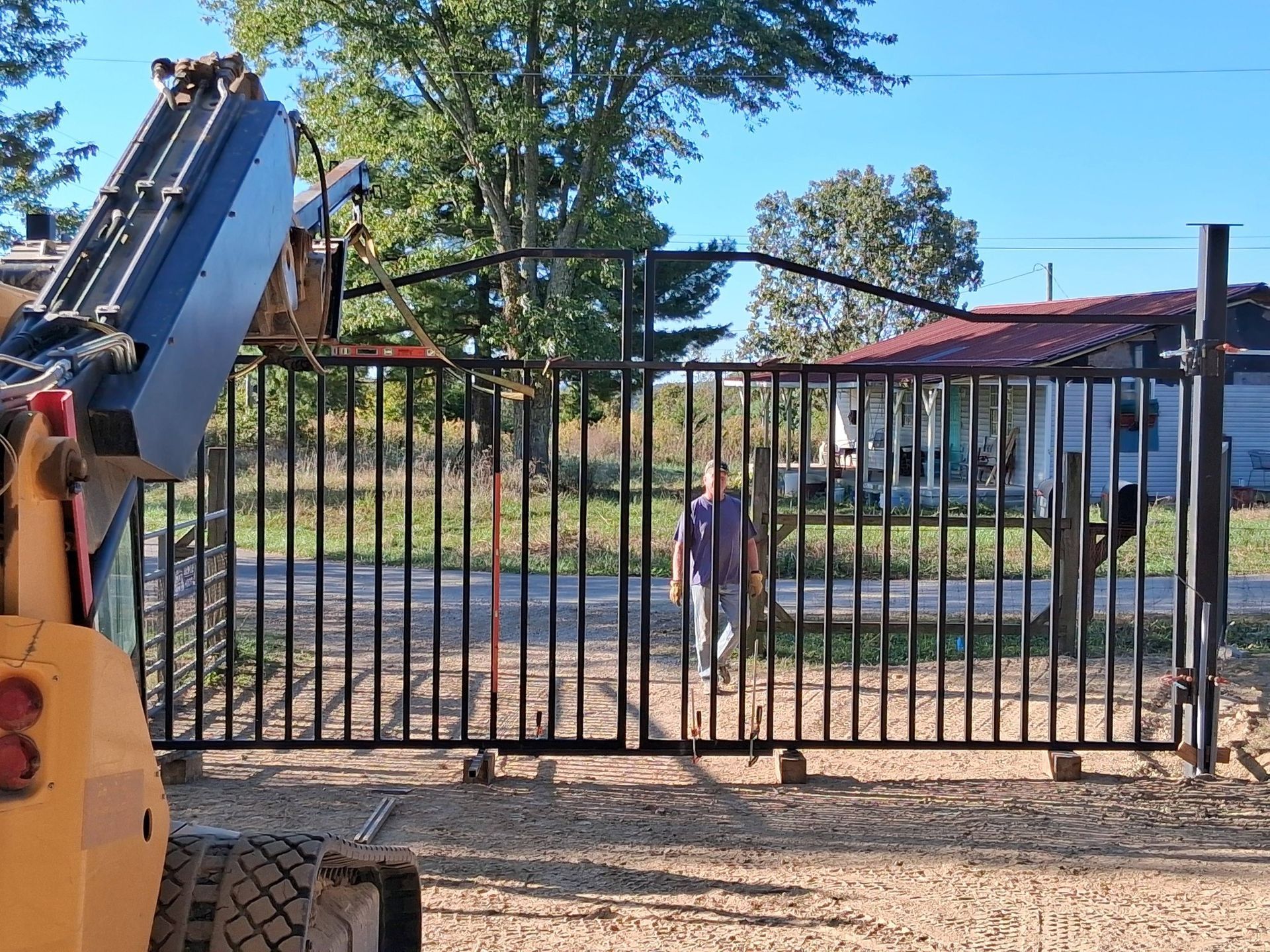 Black metal gate being installed, person standing in the opening; a small bulldozer is in the foreground.