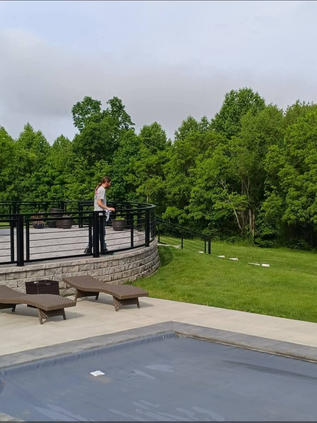 Person standing on a patio overlooking a pool and green trees on an overcast day.