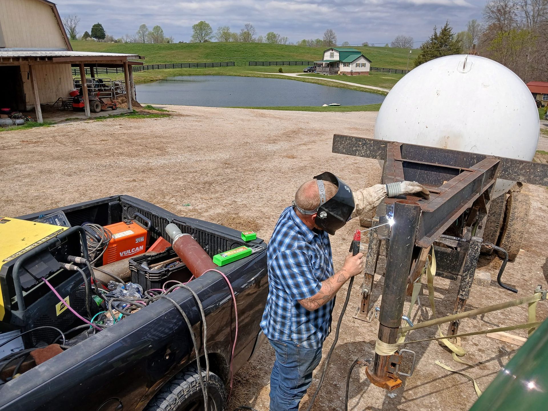 Man welding on a trailer next to a truck. A white dome and pond are in the background.
