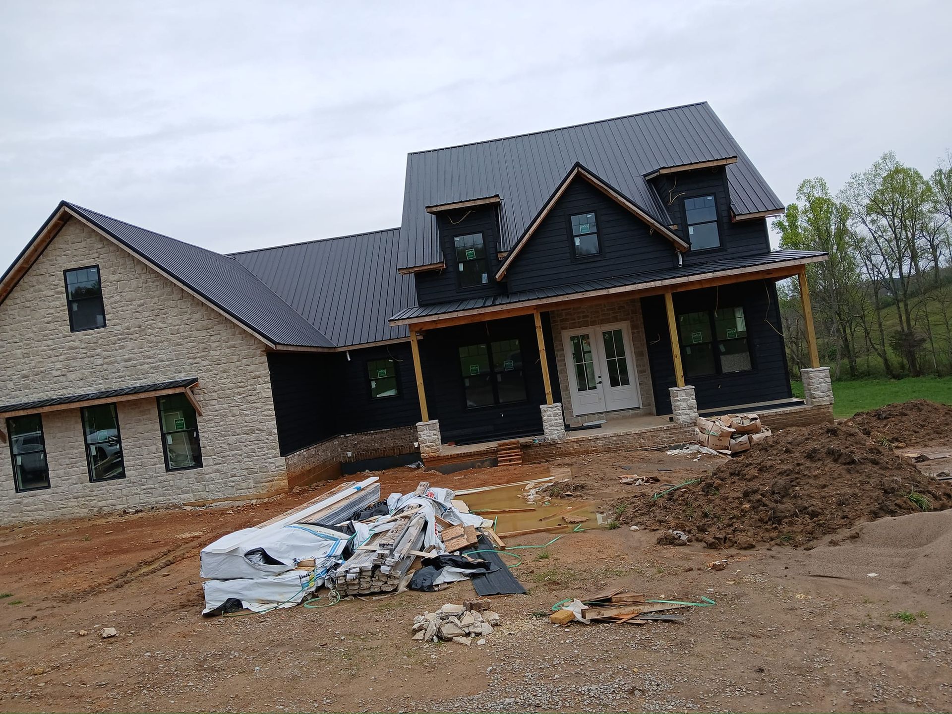 House under construction with stone and black siding, black metal roof, and scattered construction materials.