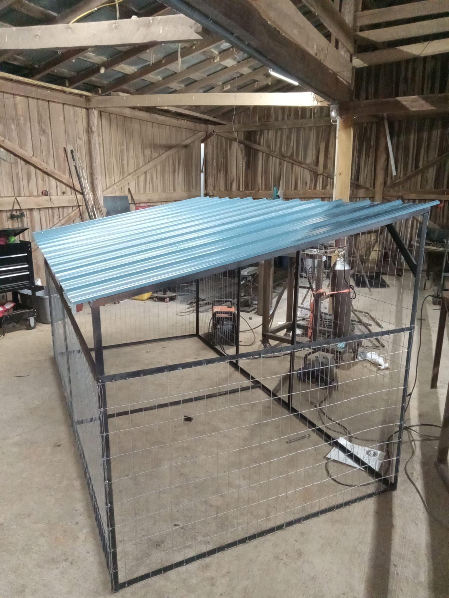 Metal-framed outdoor enclosure with a blue corrugated metal roof inside a barn.