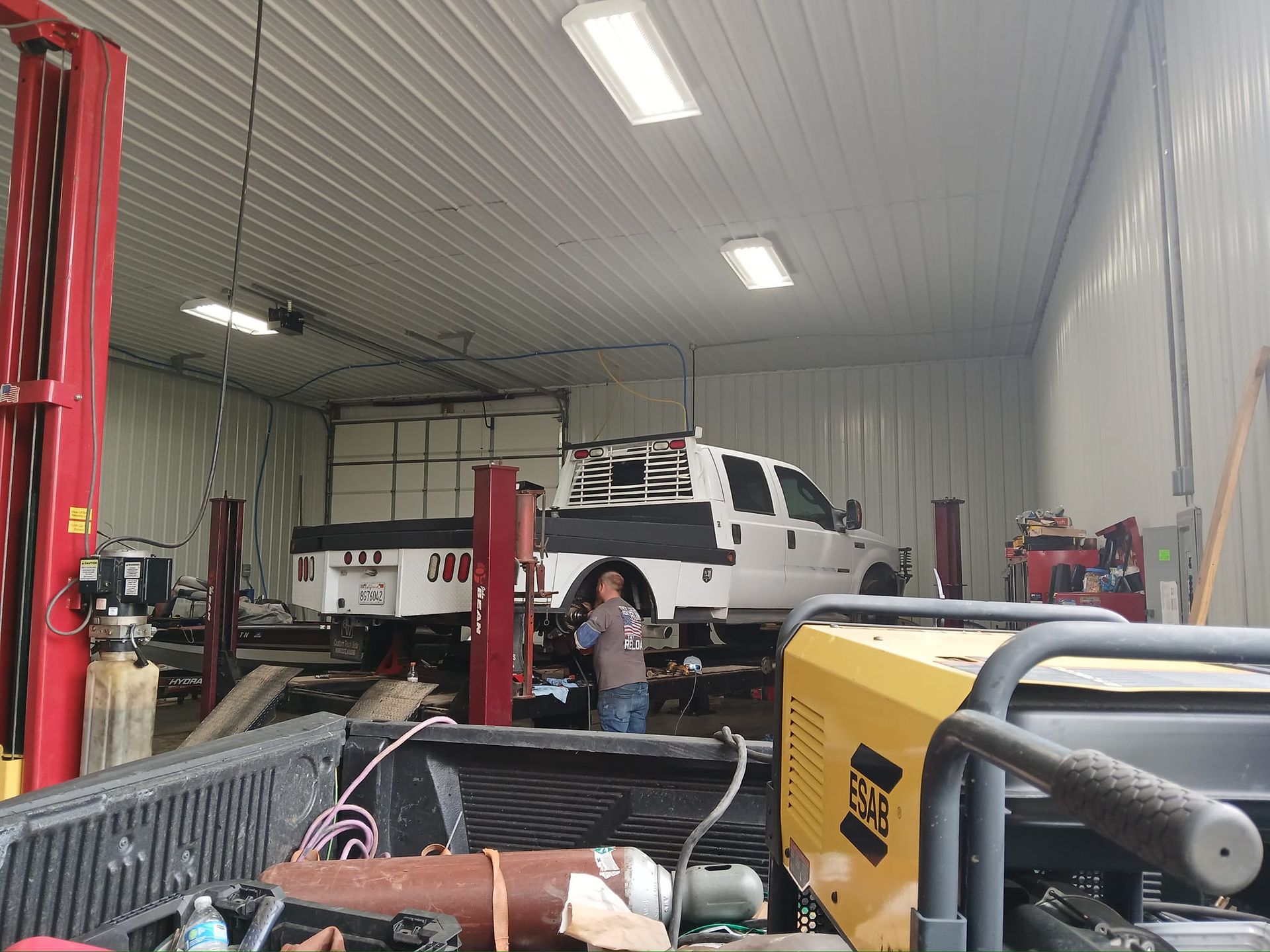 Inside a repair shop, a white truck is raised on a lift; a person stands nearby.