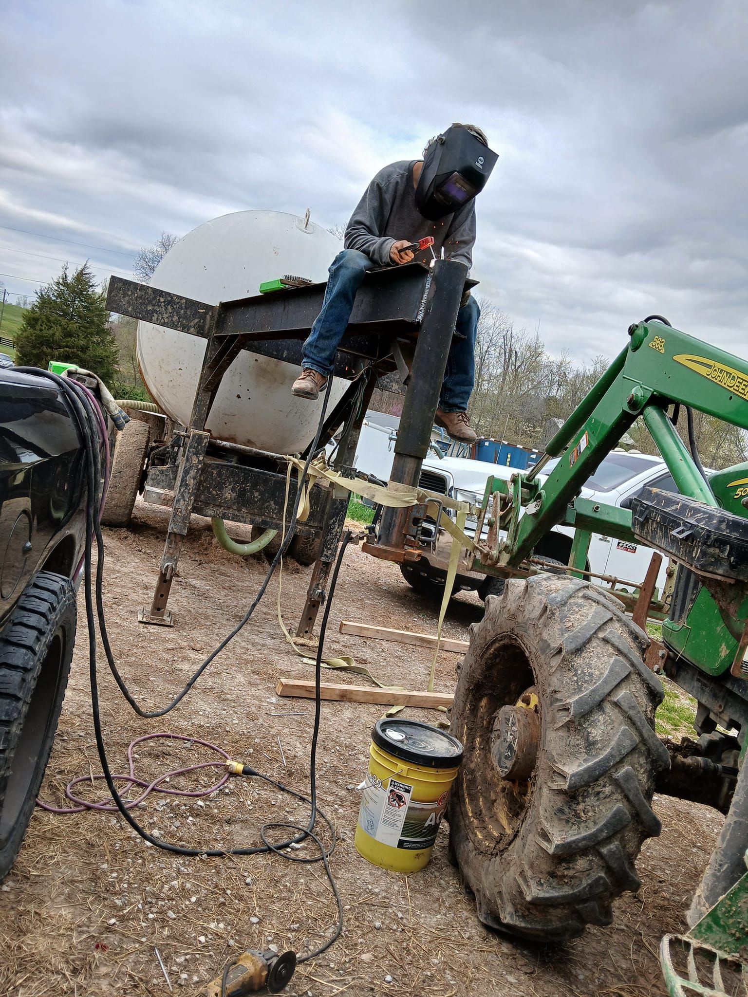 Person welding atop a large farm implement, wearing a welding mask. Outdoors, John Deere tractor, cloudy sky.