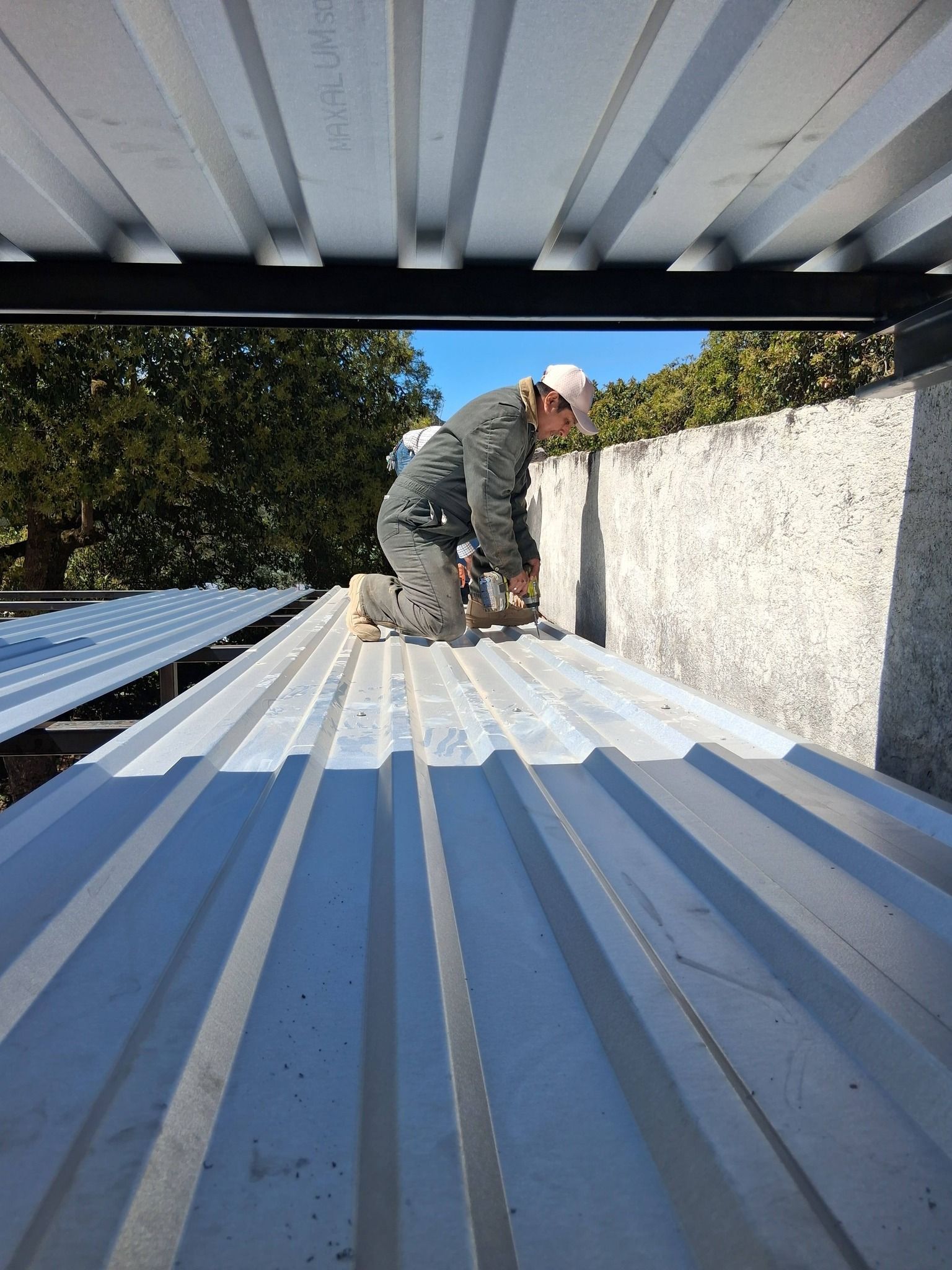 A person in a hard hat kneeling on a corrugated metal roof, working on a construction site.