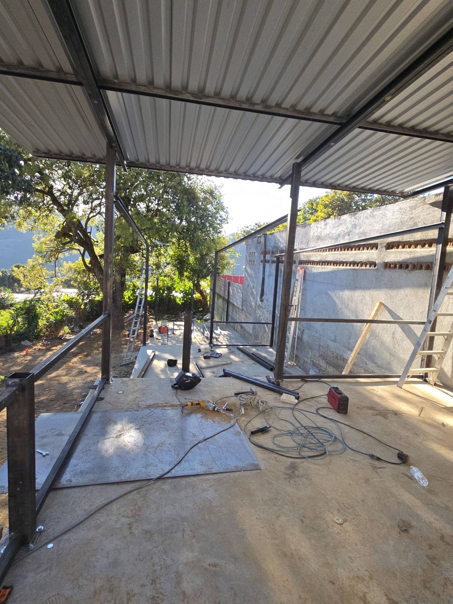 Construction site under corrugated metal roof. Steel framework, welding tools, unfinished walls, and foliage visible.