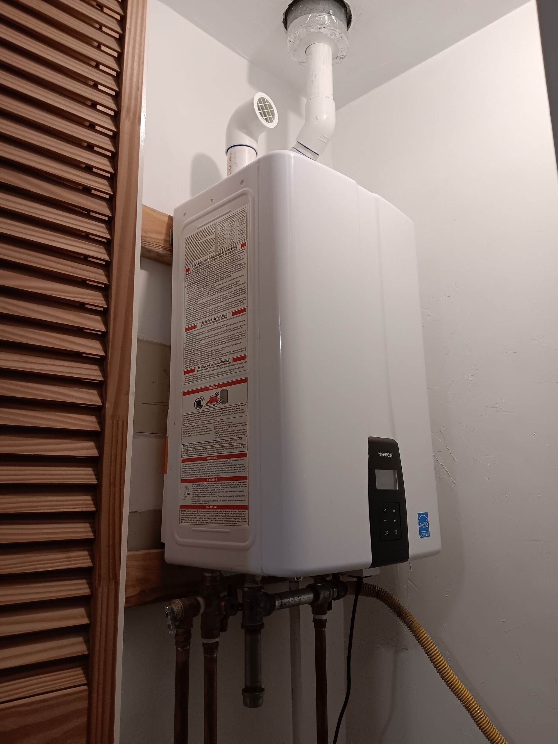 Wall-mounted white water heater with pipes and venting in a narrow indoor utility space