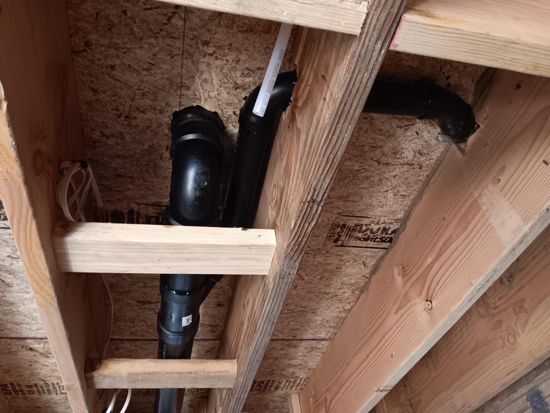 Black PVC plumbing pipes and wooden joists inside a crawlspace ceiling.