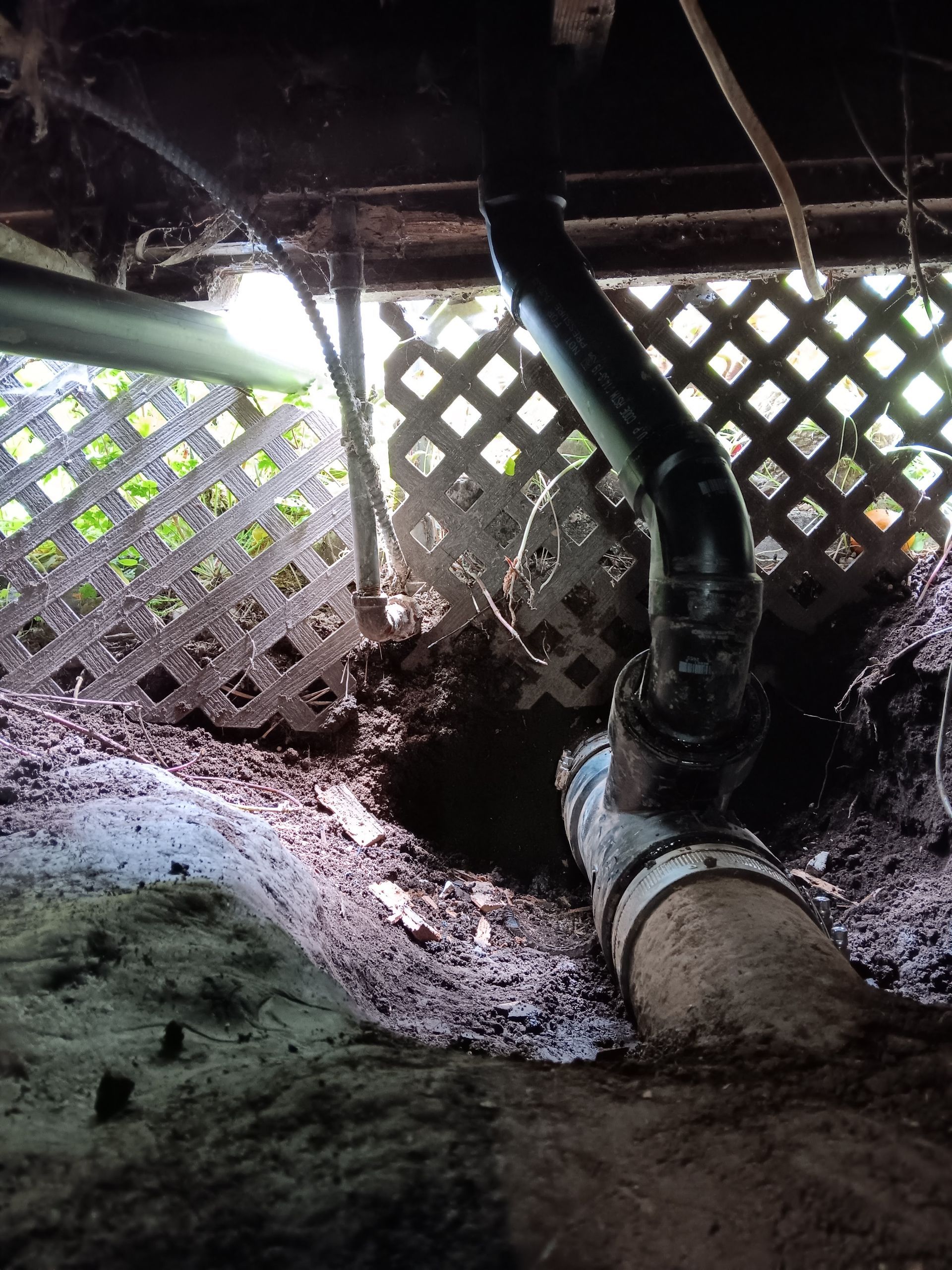 Crawling dark pipe under a lattice-covered crawlspace, with muddy ground and daylight filtering through.