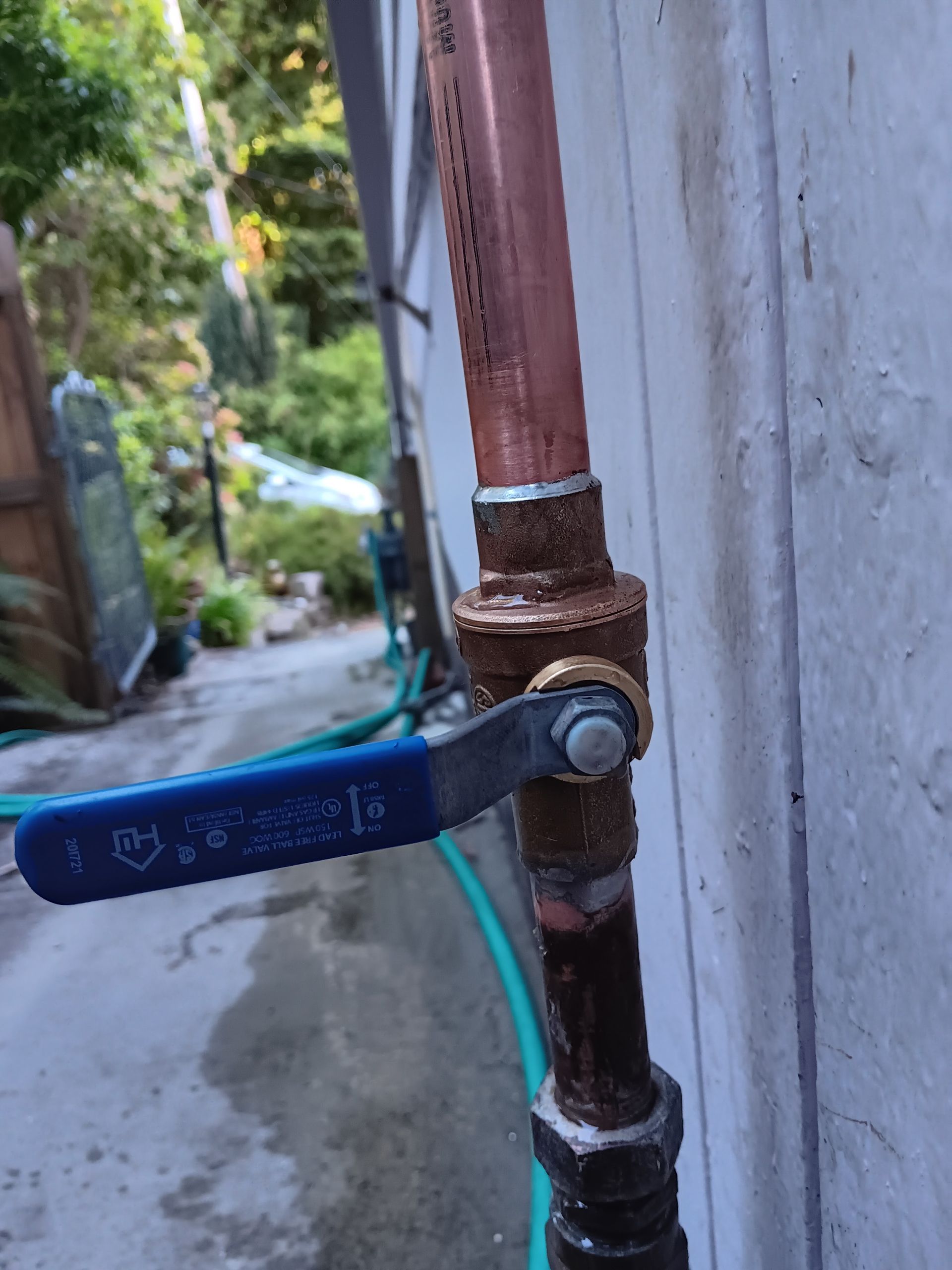 Blue-handled water shutoff valve on a rusted pipe beside a wall in a narrow alley.