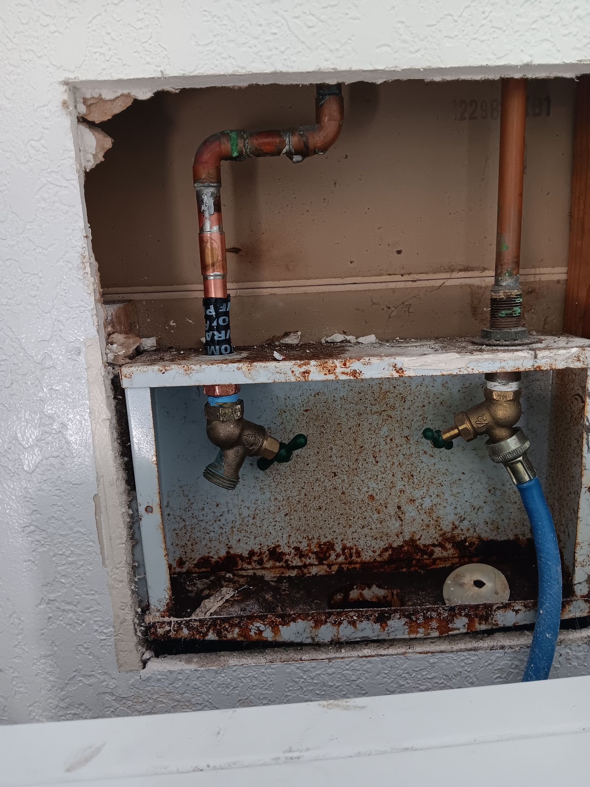Open wall cavity with old copper pipes, rusted metal box, and blue hose connected to valves