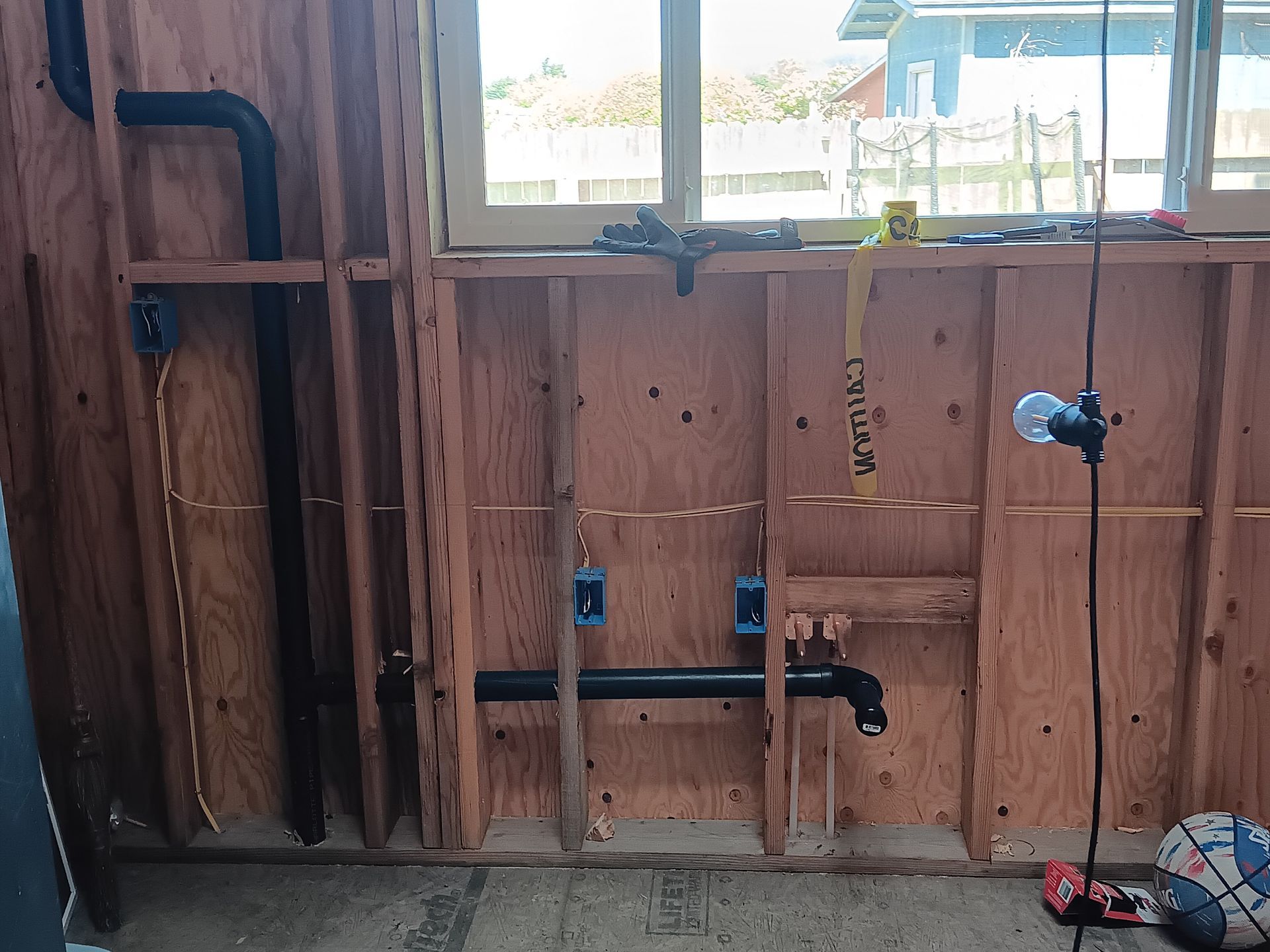 Rough plumbing wall with exposed pipes, valves, and a window above in an unfinished room.