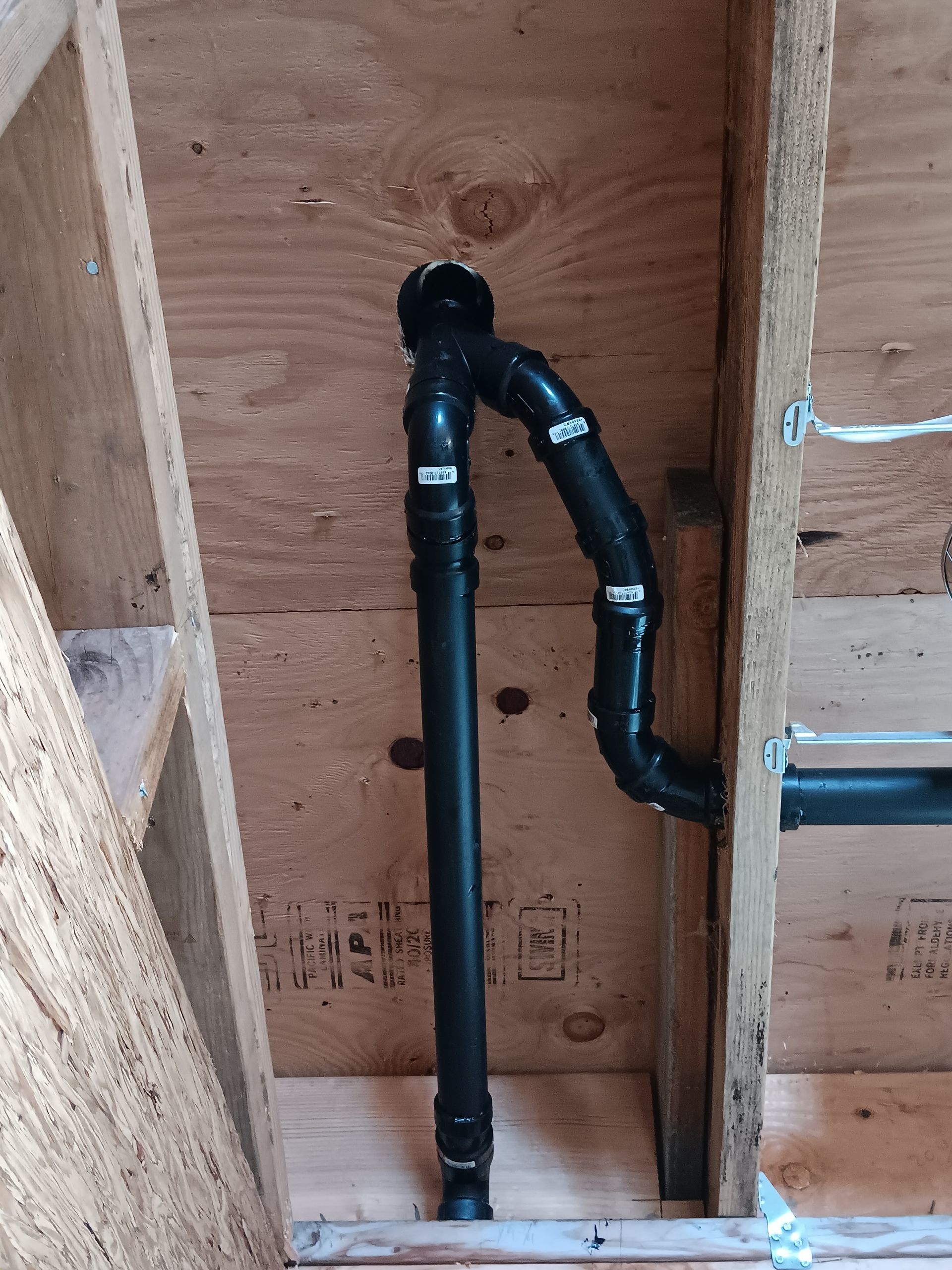 Black PVC pipes run vertically and bend inside an unfinished wooden wall cavity.