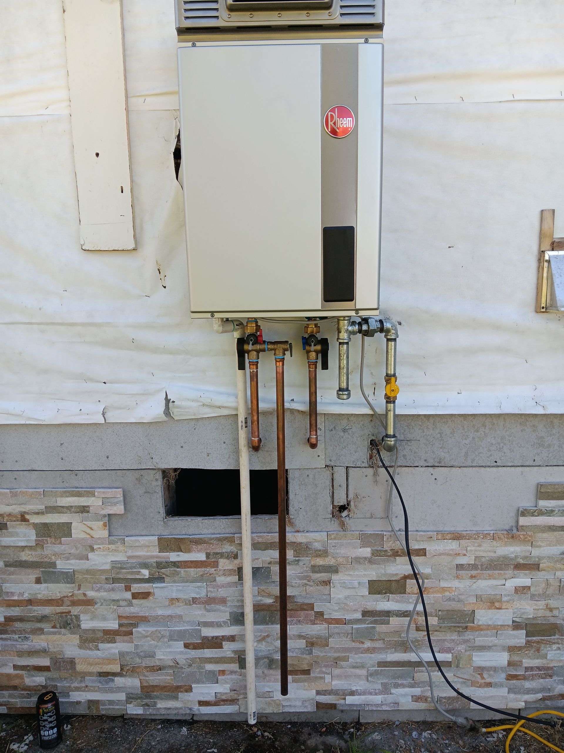 Outdoor wall-mounted utility unit with exposed pipes and wiring above a stone foundation