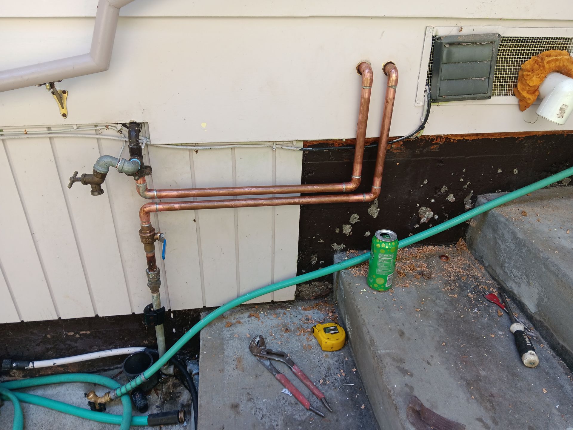 Copper pipes and a valve mounted on an exterior wall beside a concrete step, with tools and a green hose nearby