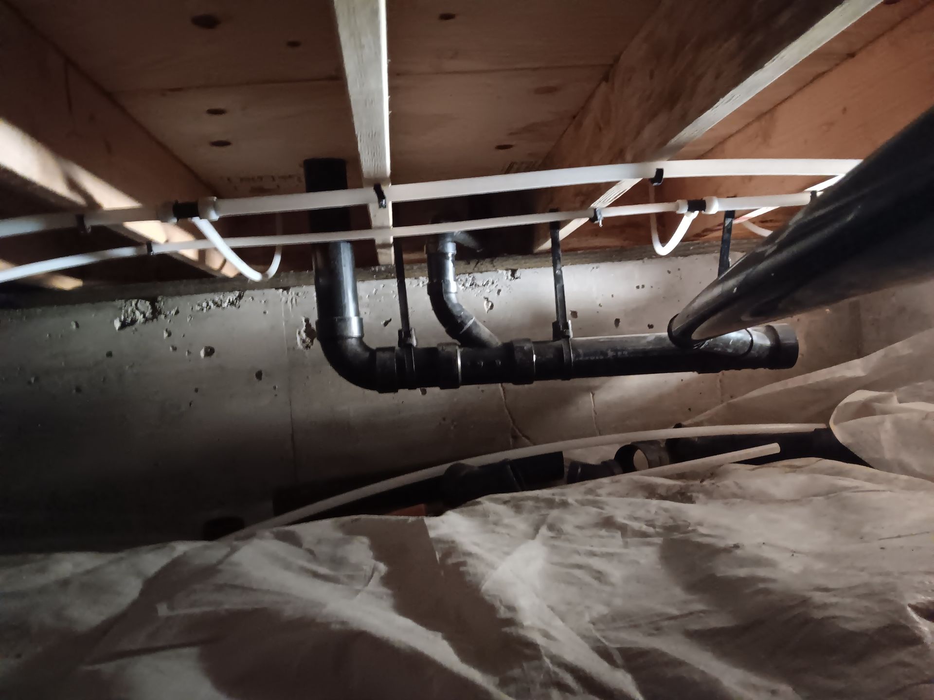 Exposed black plumbing pipes beneath floor joists in a dim crawlspace, with insulation in the foreground