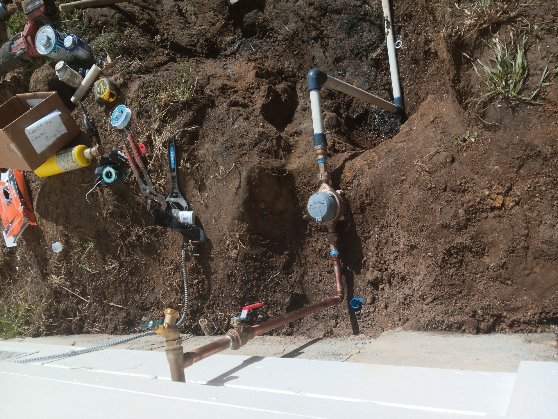 Exposed water pipes and fittings in a dirt trench beside a concrete wall during utility work