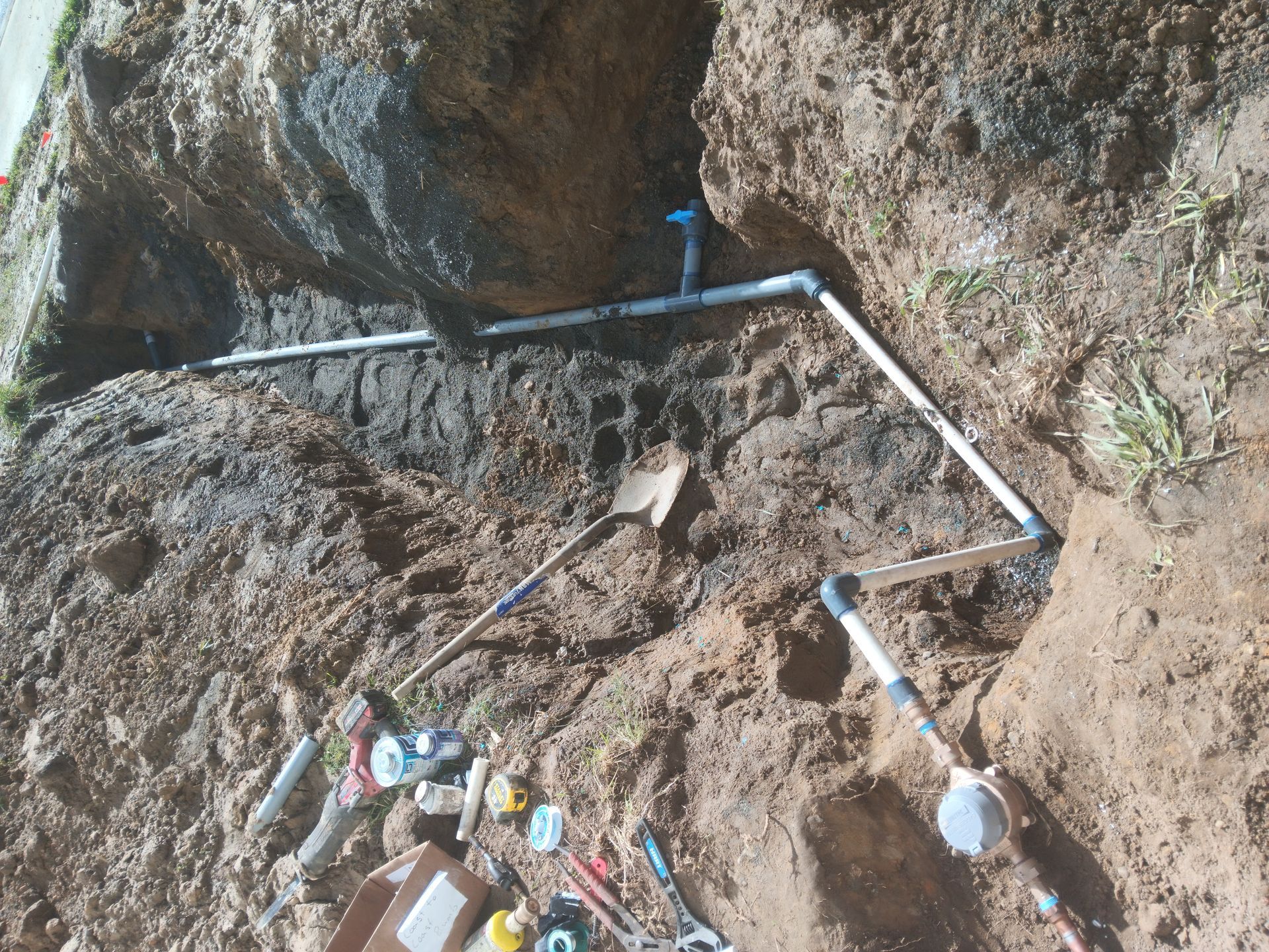 Rocky slope with white PVC pipes and a few tools near a muddy trench.