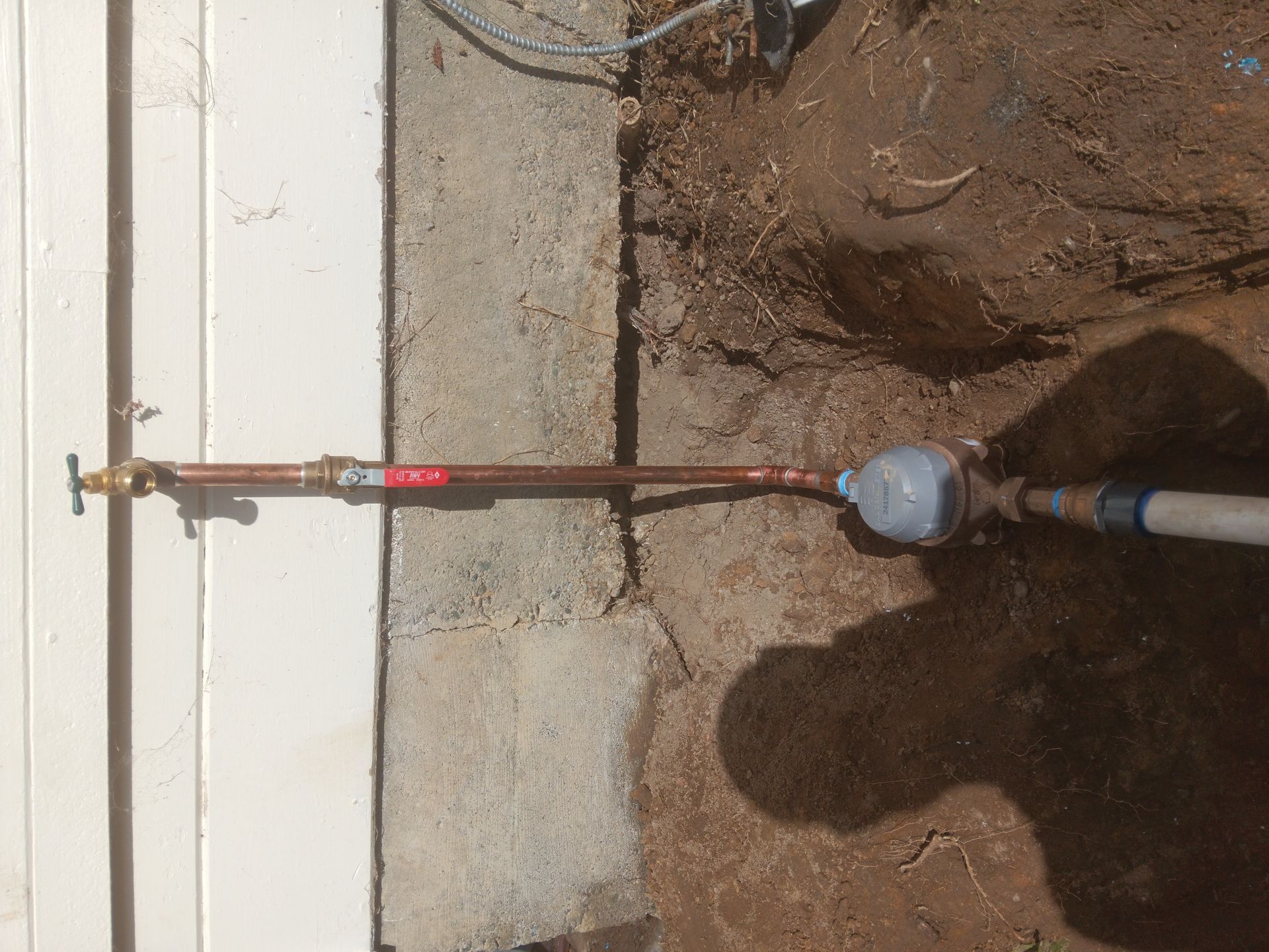 Pipe wrench connected to a red pipe section near a wall, outdoors over dirt and concrete.