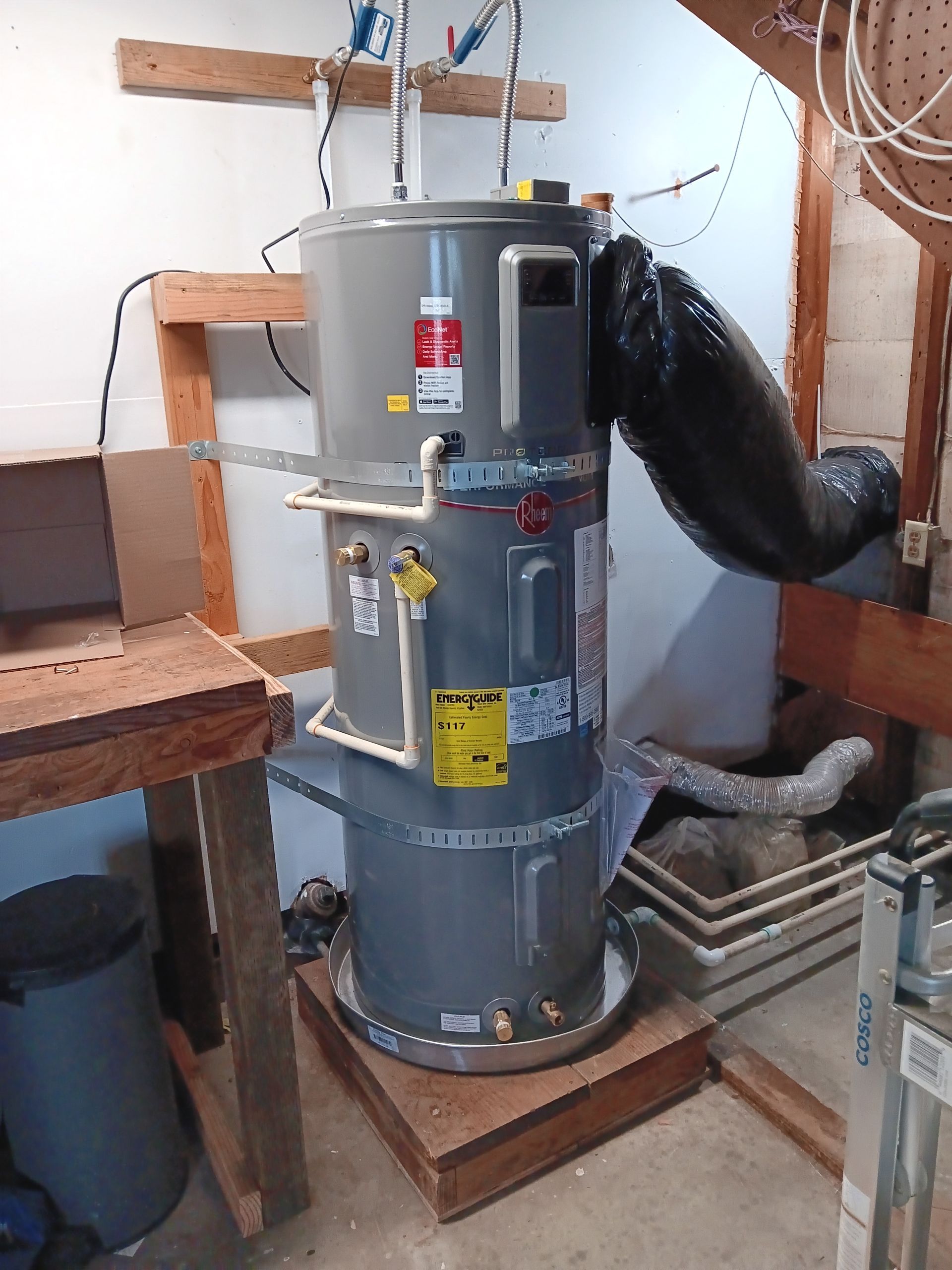 Gray water heater in a basement with black vent hose and wooden stairs nearby.