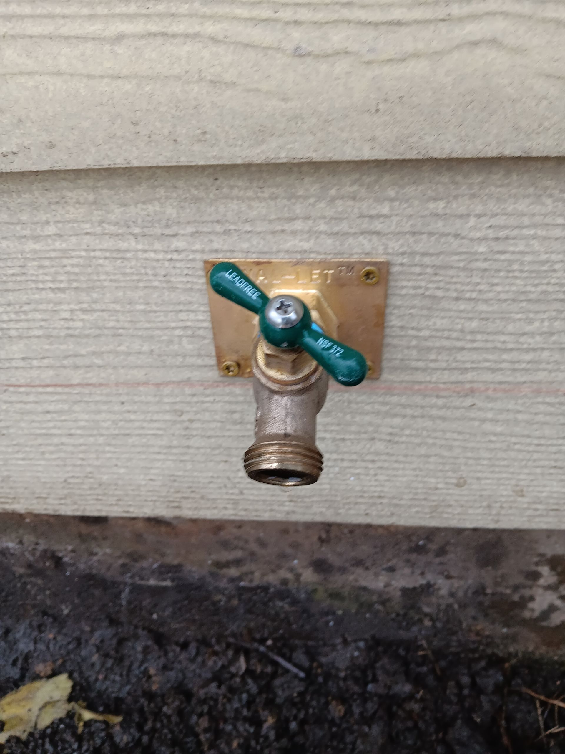 Green hose bib valve on a wooden wall above dark ground.