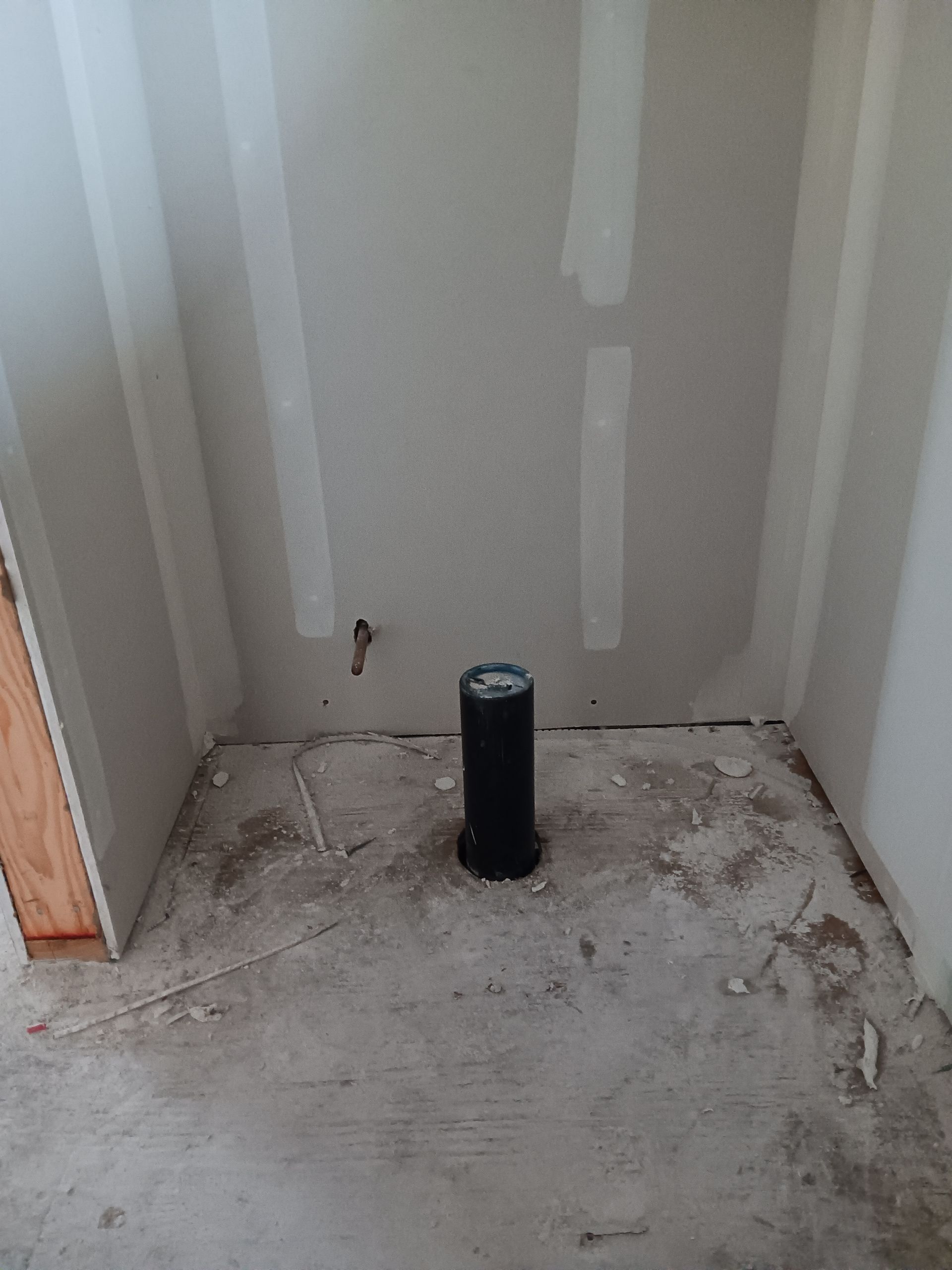Small unfinished room corner with gray drywall, patchy concrete floor, and a black pipe in the center.