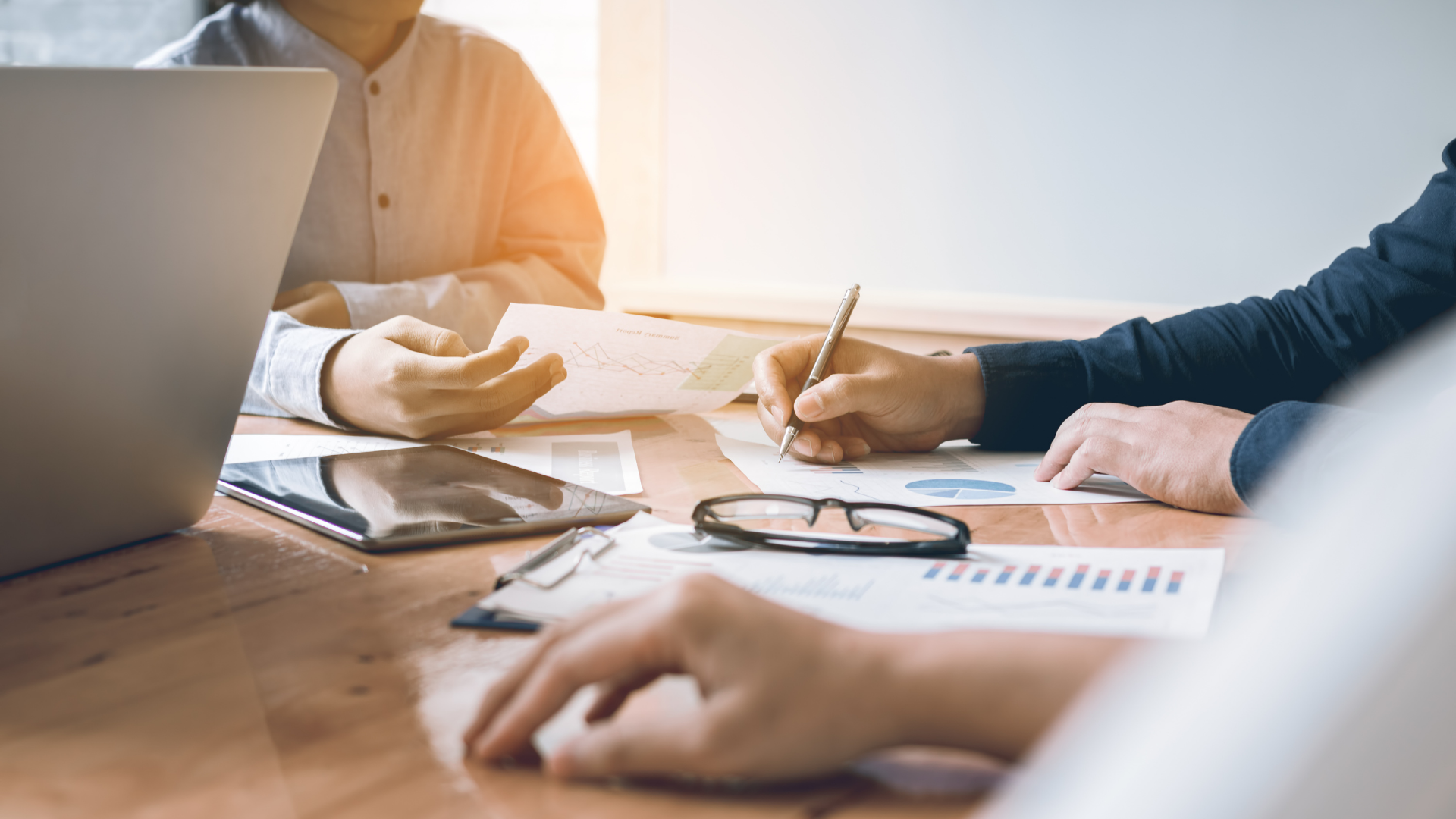 Le persone sedute a un tavolo esaminano i documenti, con un computer portatile e un tablet presenti. Una persona scrive con una penna.