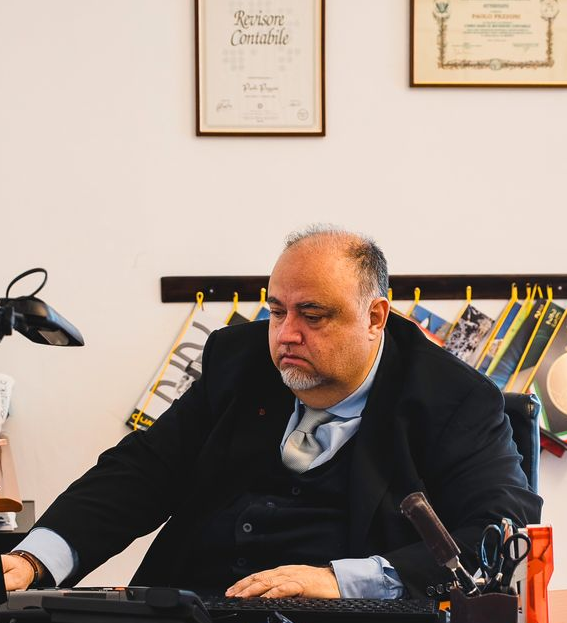 Due uomini in giacca e cravatta, uno seduto a una scrivania, l'altro in piedi, in un ufficio con documenti e una finestra.