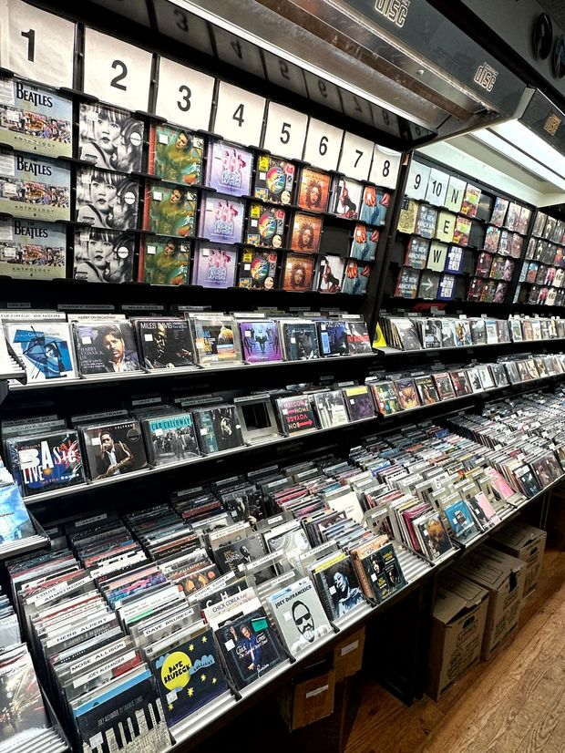 Stacks of blue Blu-ray discs on a table in front of shelves of DVDs, inside a store.