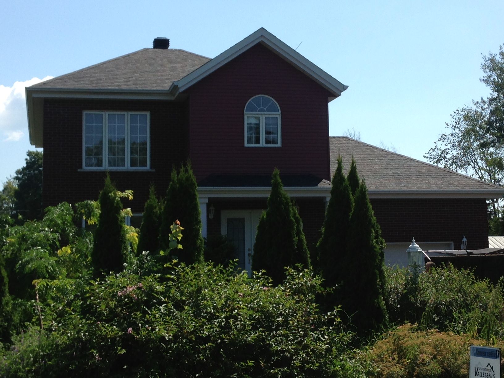 A red house with a gray roof is surrounded by bushes and trees