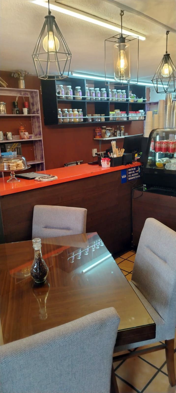 Interior de una cafetería con mesas, sillas, mostrador y estanterías. Luces decorativas cuelgan del techo.