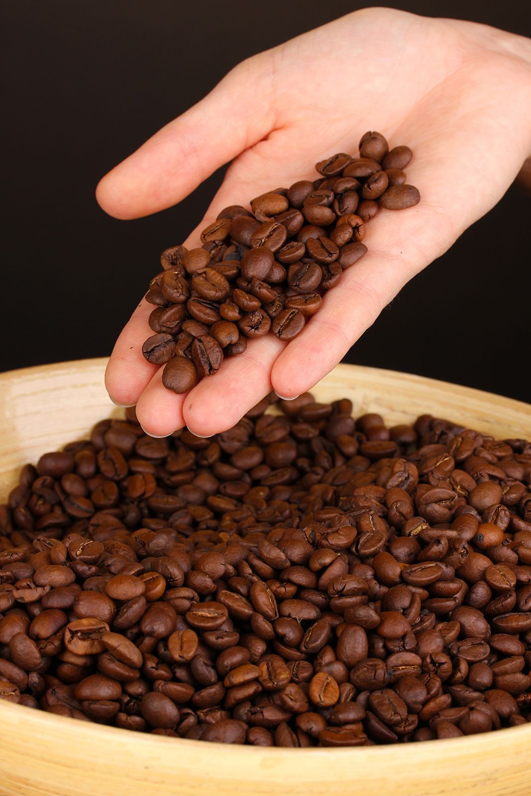 Puñado de granos de café tostados sobre un cuenco de madera lleno de granos de café; fondo oscuro.