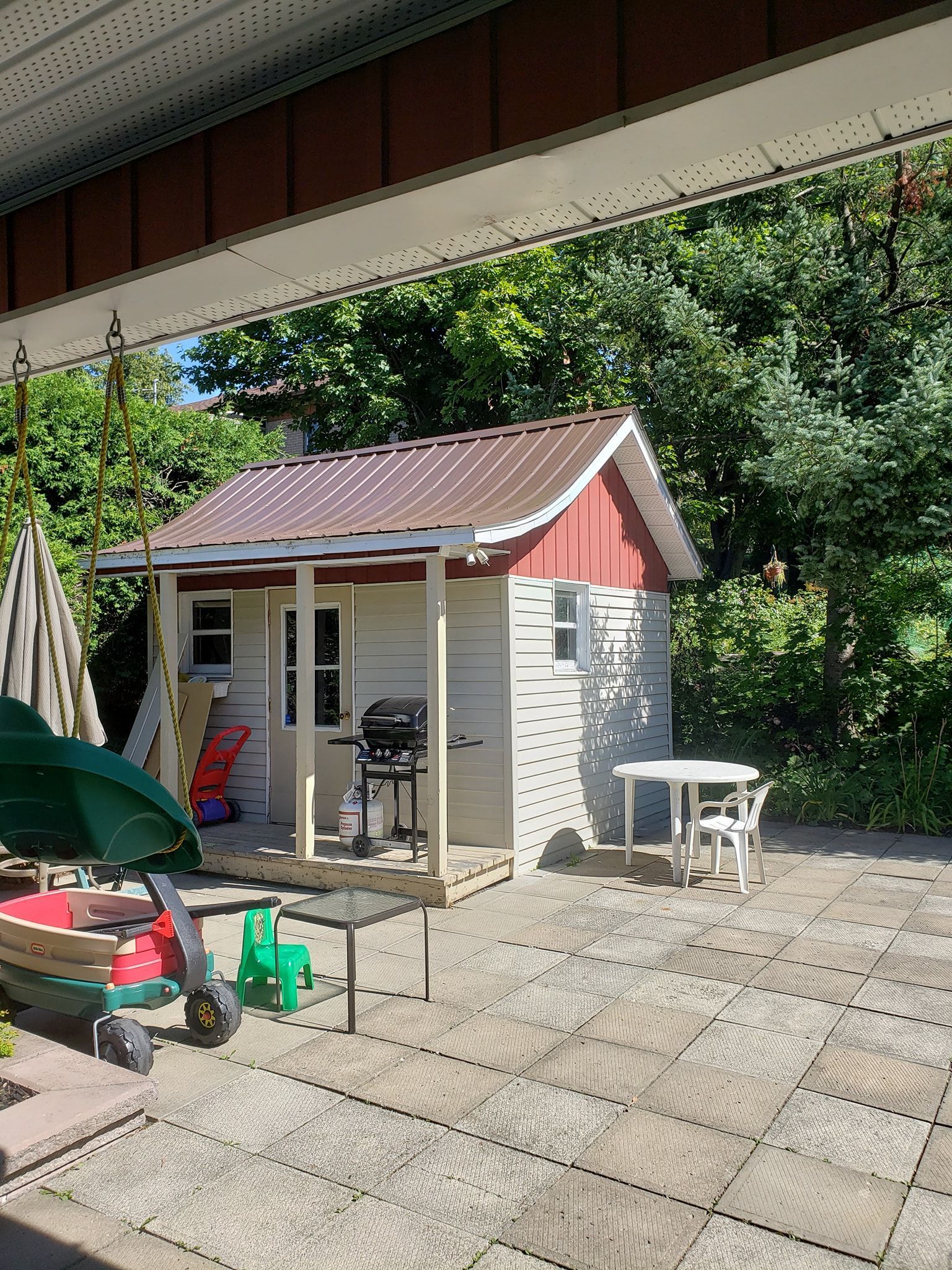 Un petit hangar blanc avec un toit rouge se trouve sur un patio à côté d'une table et de chaises.