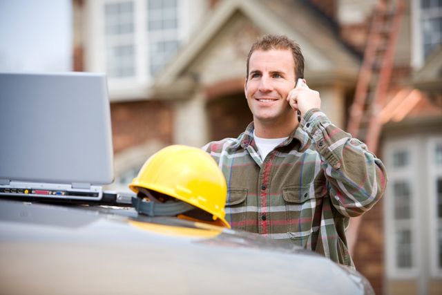 A man is talking on a cell phone while standing next to a hard hat and a laptop.