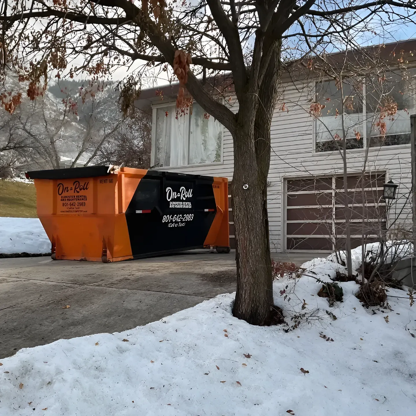 Orange and black dumpster in driveway next to a two-story brick house covered in snow.