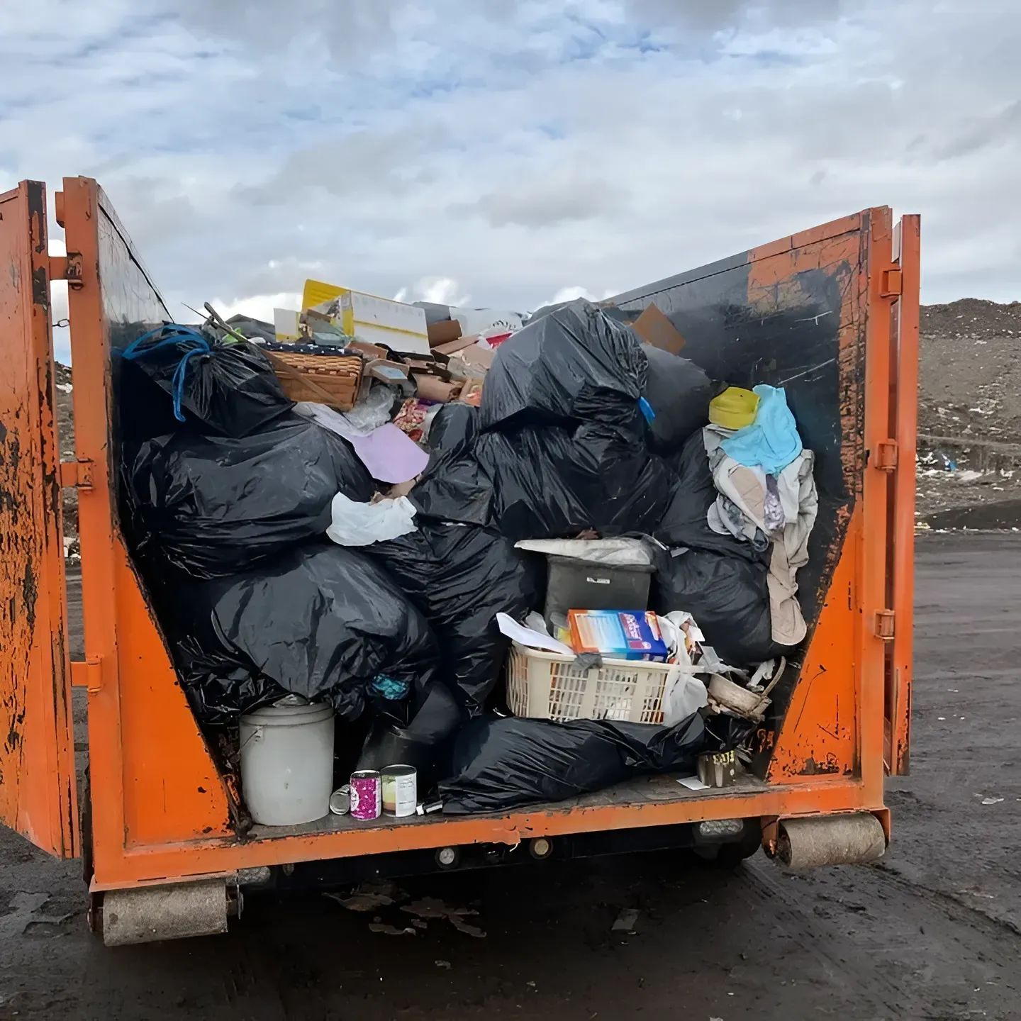 Orange dumpster overflowing with black trash bags and debris, outdoors under a cloudy sky.