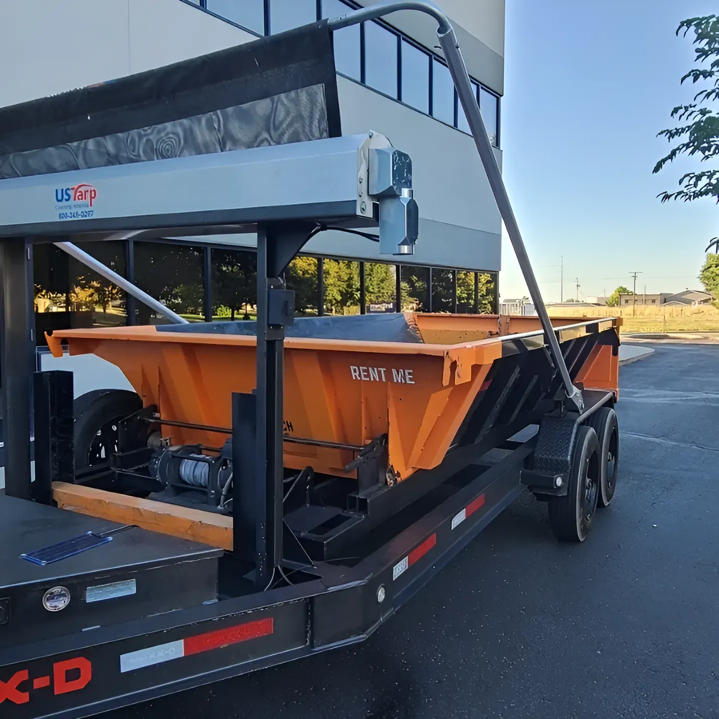 Orange asphalt distributor on a black trailer, parked outside a building.