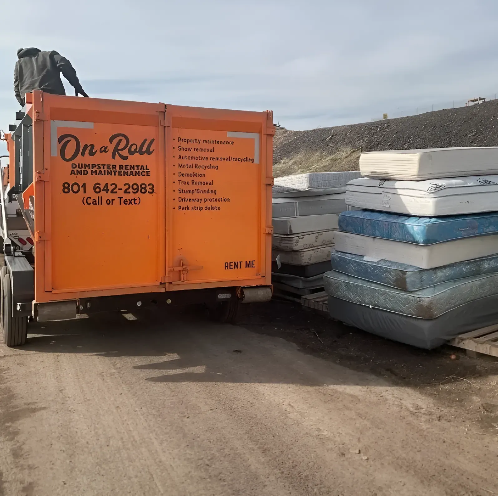 Orange On-a-Roll dumpster being loaded with mattresses by a person, outdoors on a dirt road.