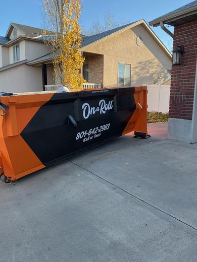 Orange and black roll-off dumpster in a residential driveway next to a brick wall and a house.