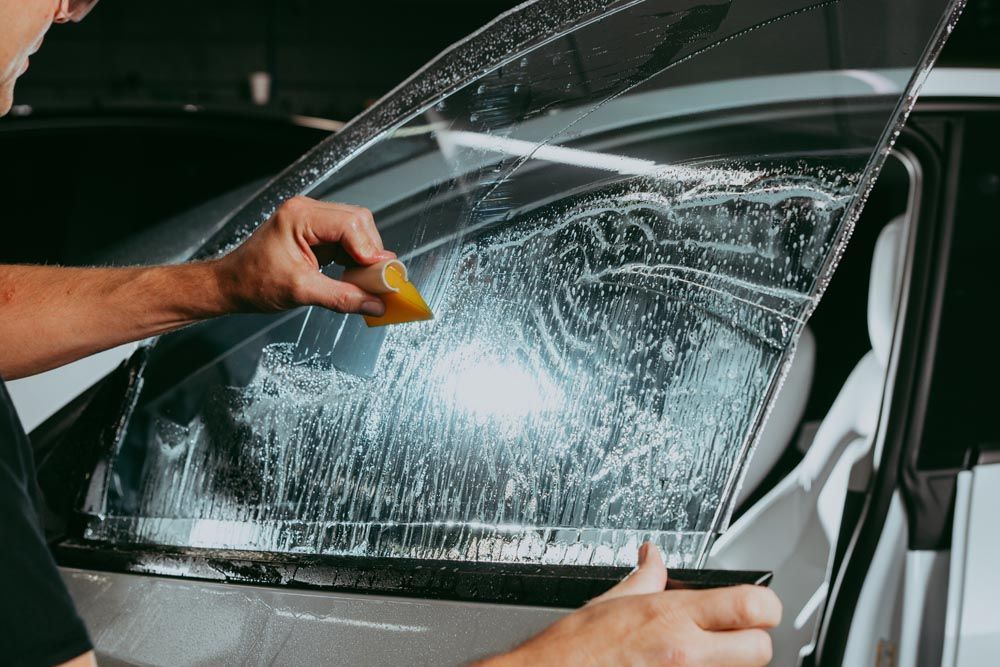 Hands cleaning a car window with a yellow scraper and cloth, covered in soap suds.
