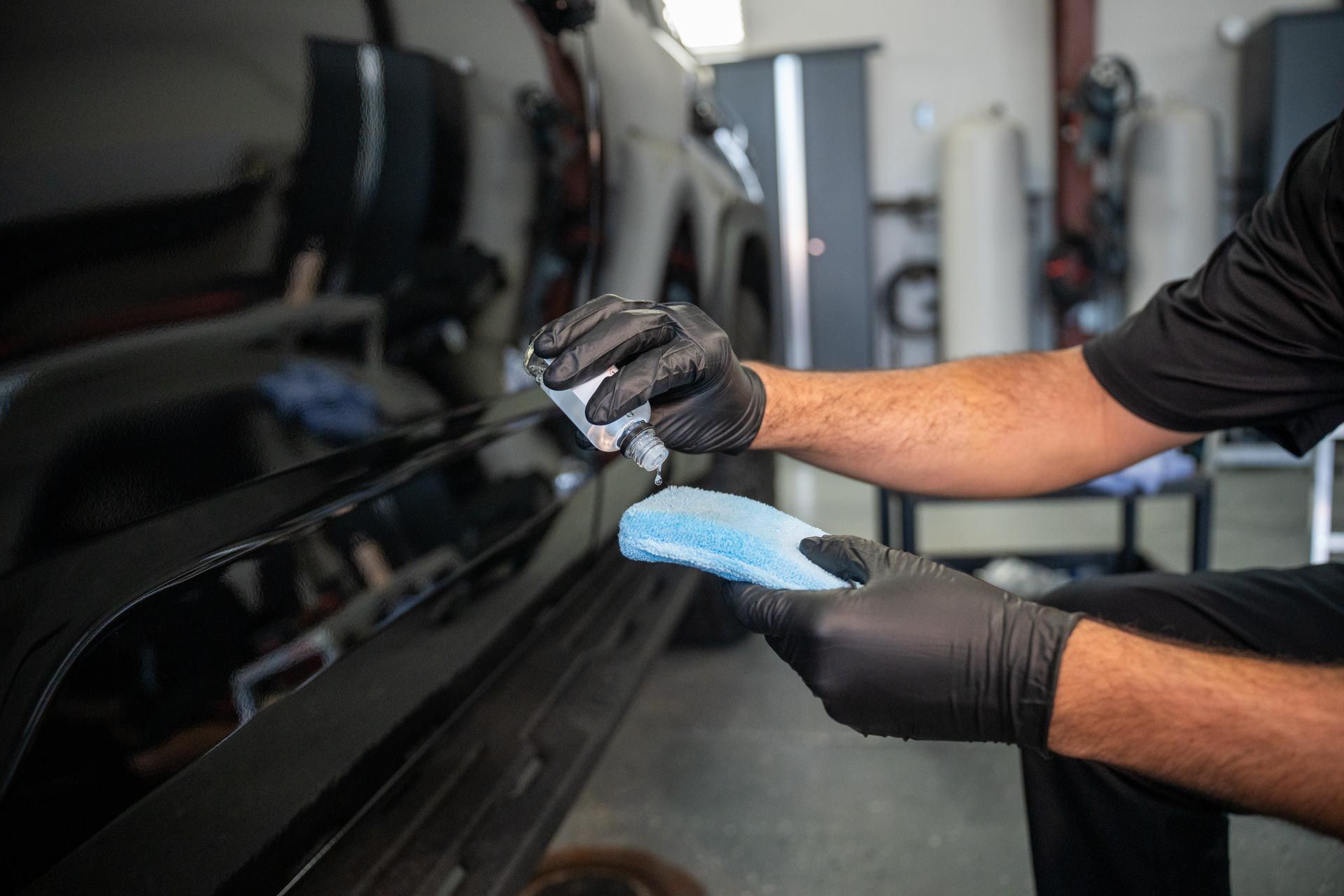 Hands in black gloves polishing a black car with a blue cloth in a garage.