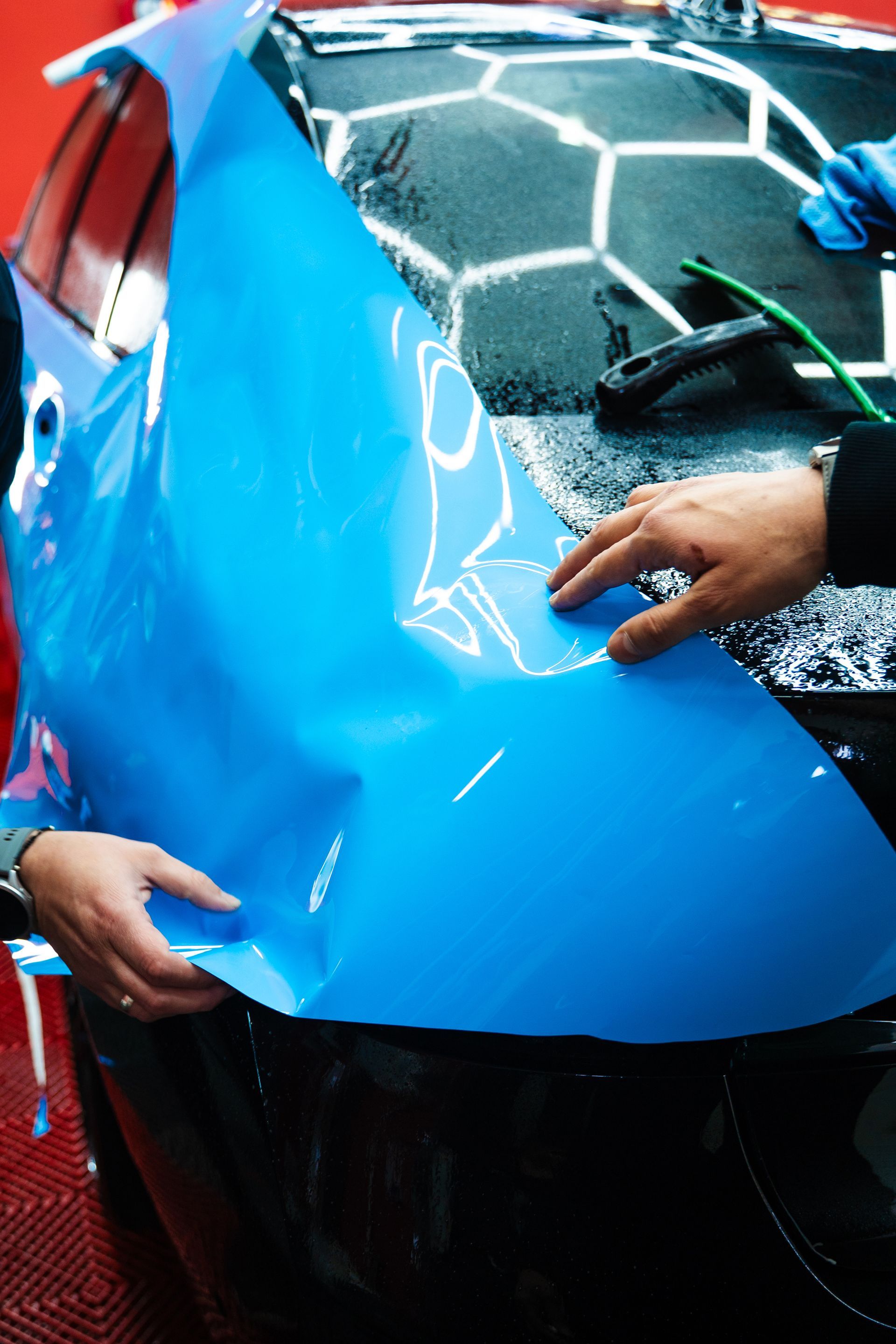 Hands applying bright blue vinyl wrap to a car hood in a workshop